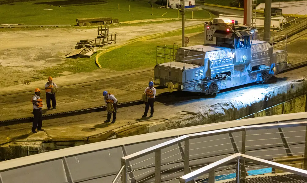 Four Panama Canal workers in hard hats and life vests hold coiled ropes beside a towing locomotive at the Gatún Locks, at 19:59