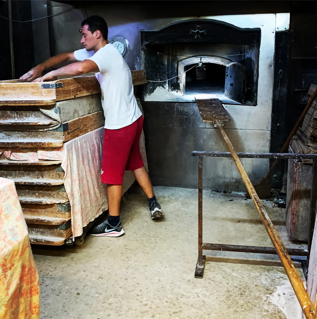 A baker in a white T-shirt slides trays of bread beside a wood-fired oven in Valletta at 01:27